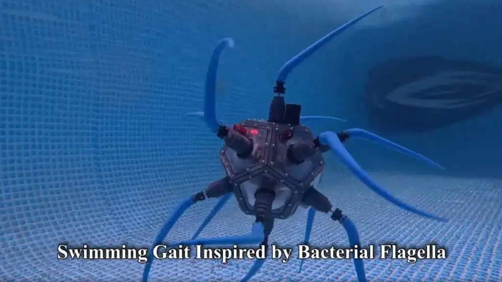 A 12-sided marine exploration drone swims in a blue tiled pool. Each face side has a blue tentacle, allowing it to swim. On-screen text reads 'Swimming Gait Inspired by Bacterial Flagella'.