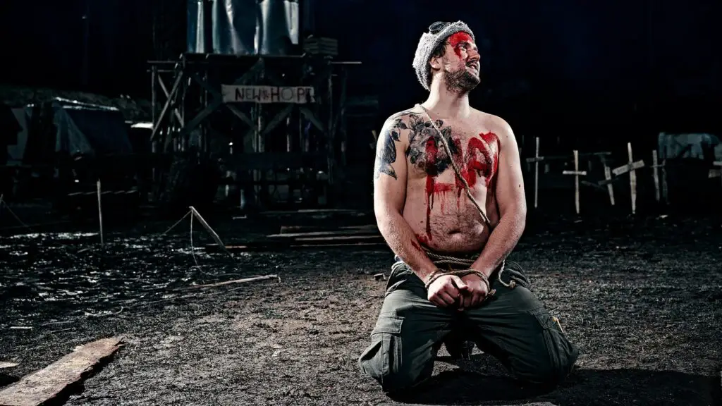 A shirtless man kneels, handcuffed, with the word 'LIAR' written in red paint across his chest.