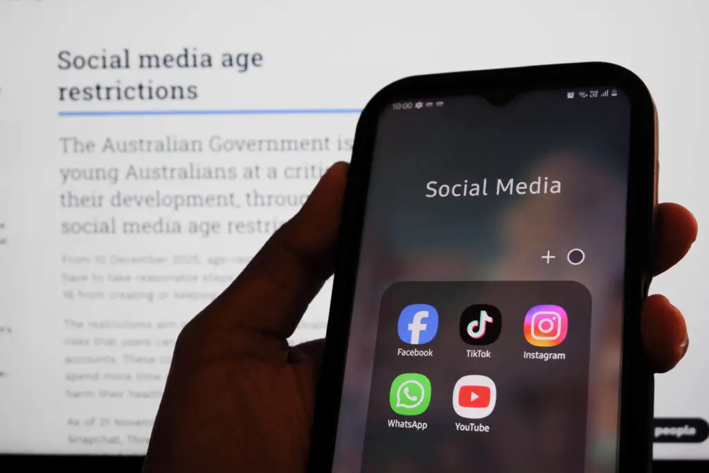 A mobile phone showing social media apps in front of a screen stipulating Australia's social media age restrictions