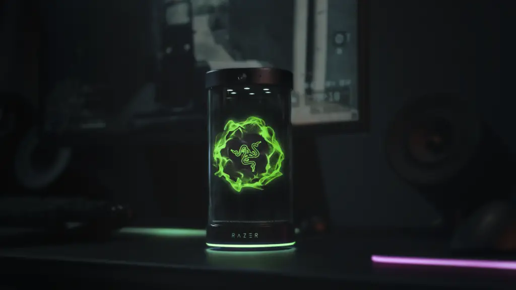 The Project AVA desk companion from Razer sits atop a black desk. A holographic image of Razer's logo lights up the screen in green.