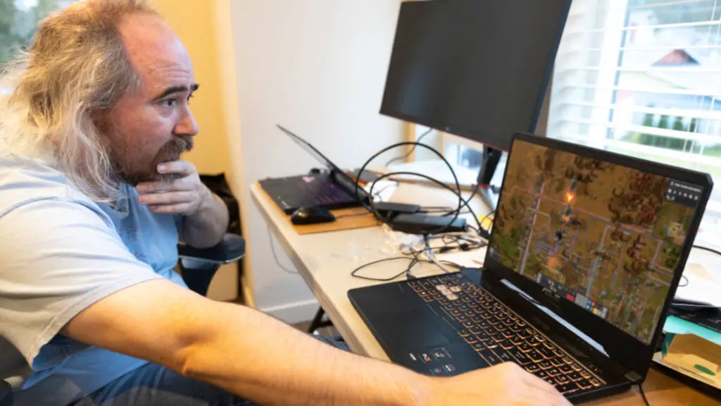 Tarn Adams sitting in front of a laptop, edited with a screen from Factorio