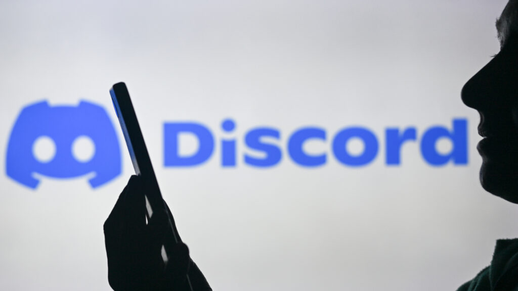 A woman holding a smartphone in front of a computer screen displaying the Discord logo.