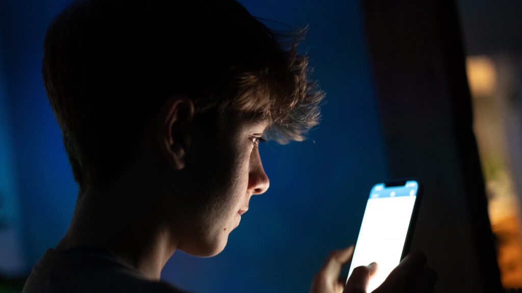A 13-year-old teenage boy looks at an iPhone screen displaying various social media apps.